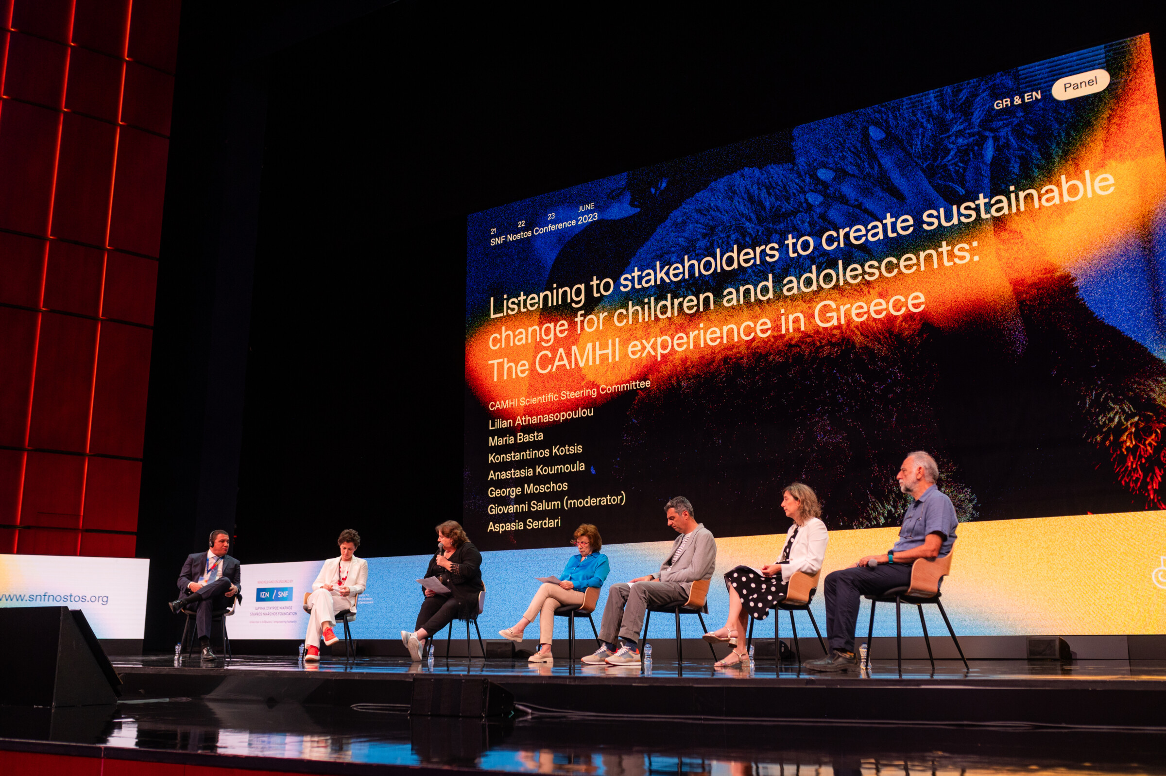 The seven speakers are seated in a row on stage, with Lillian Athanasopoulou holding the microphone.