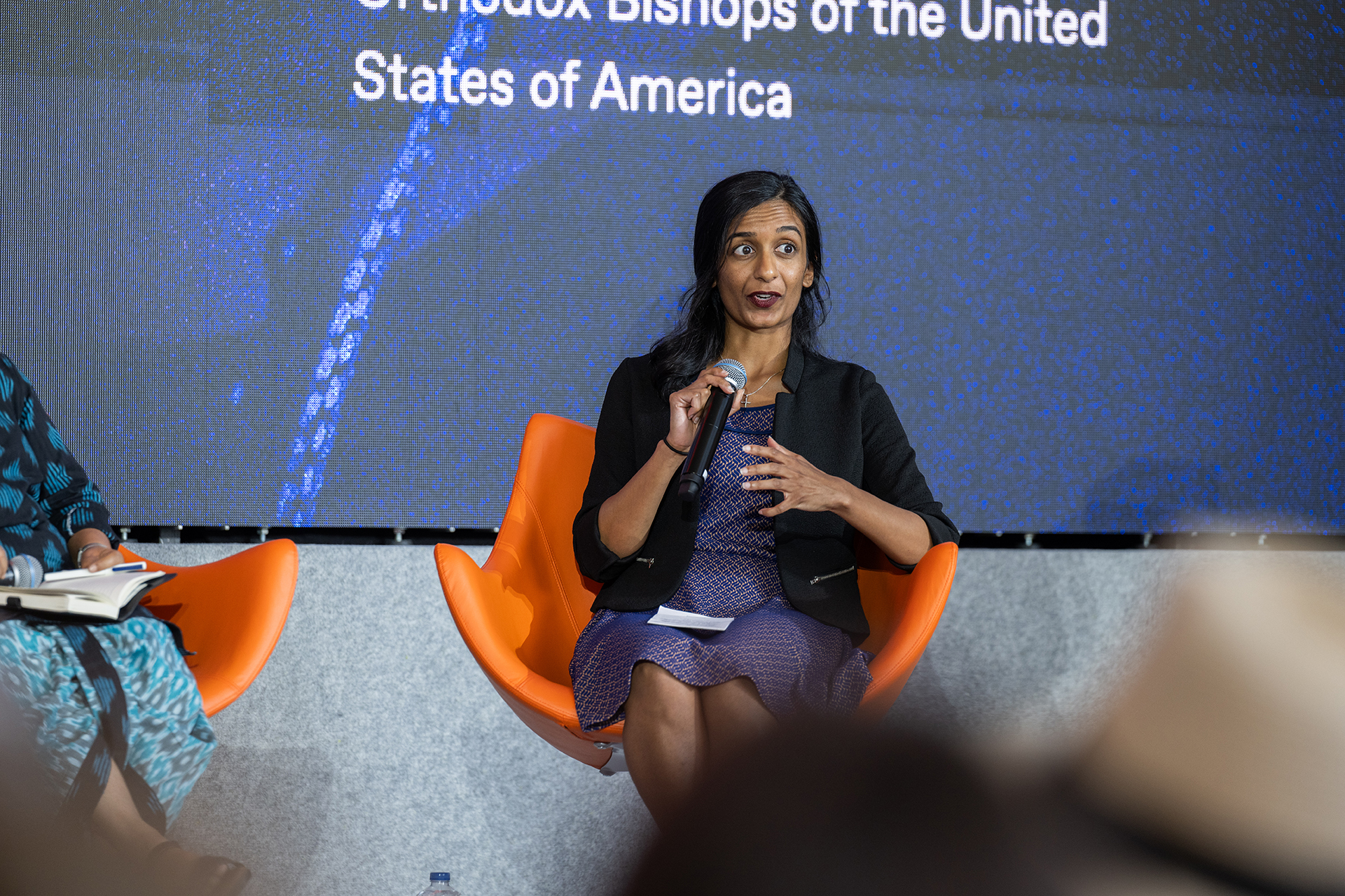 Sangeetha Thomas, wearing a purple dress and black jacket, holds the microphone and sits in an orange chair at the panel.