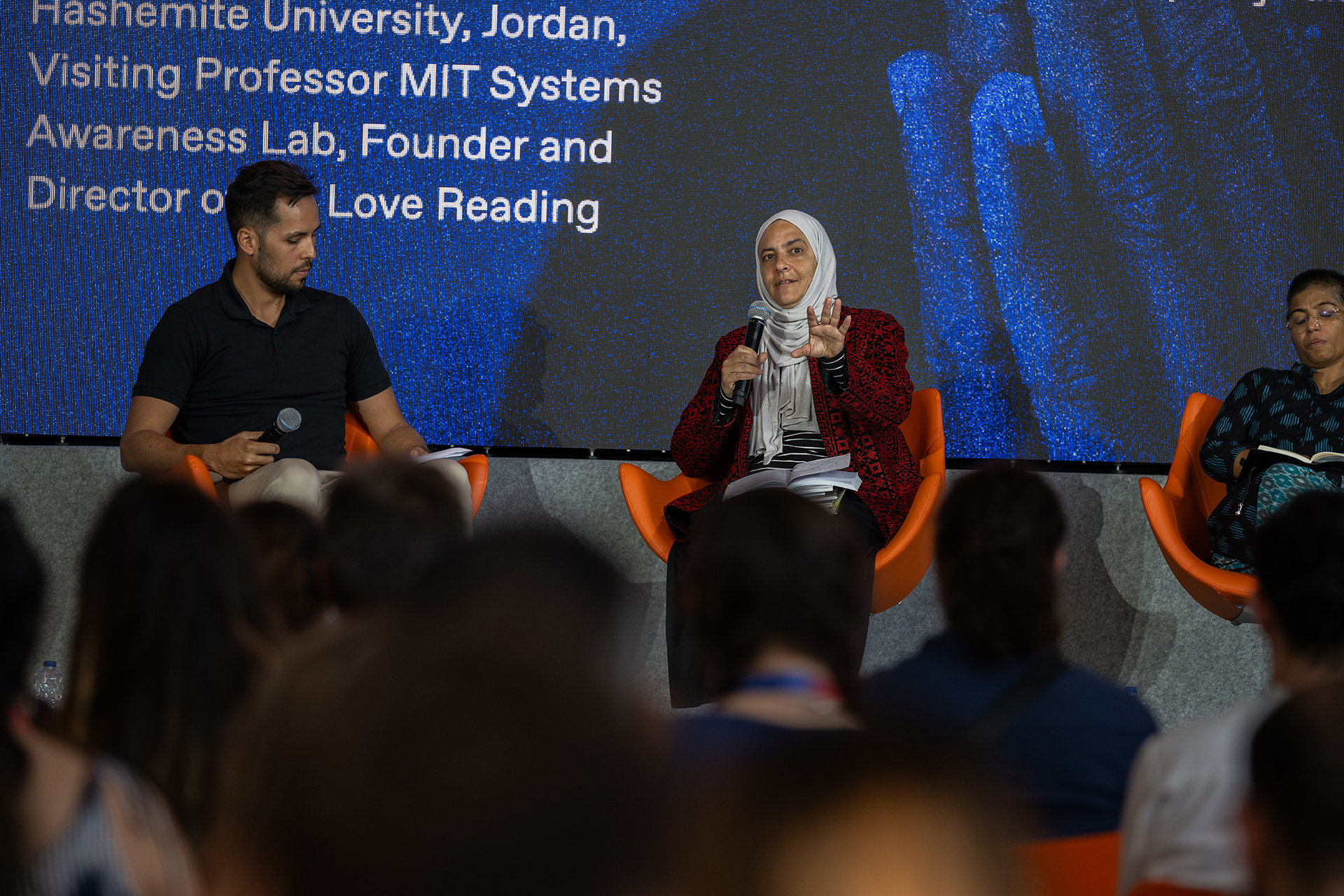 Michael Nikonchuk (left) and Rana Dajani (center) sit on the panel. The microphone is held by Rana Dajani who has her notes on her lap.