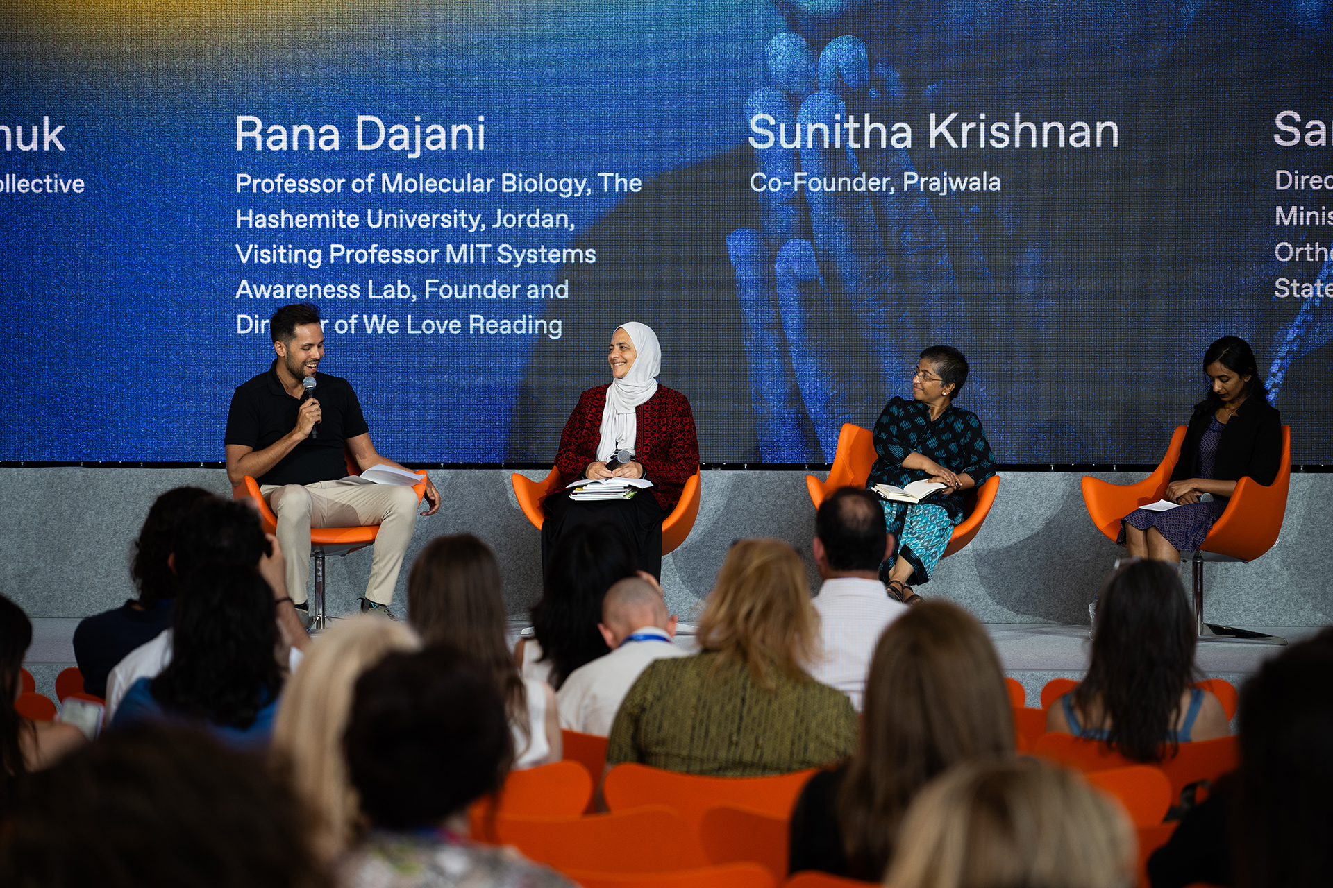 Michael Nikonchuk (left), Rana Dajani, Sunitha Krishnan and Sangeetha Thomas (right) at the Religion, Culture and Mental Health panel. They sit in orange chairs in front of a big screen, while people sit and listen to them. Michael Nikonchuk holds the microphone.
