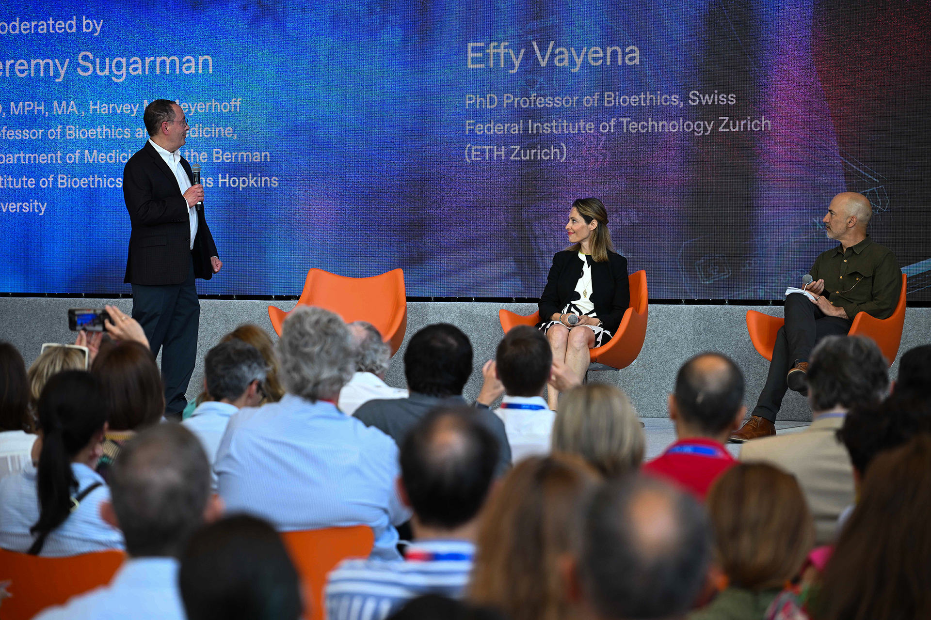 Efi Vagena is sitting between two speakers on the panel stage and looks to her left at Jeremy Sugarman who is standing and holding the microphone. Below them, the large audience is watching. 