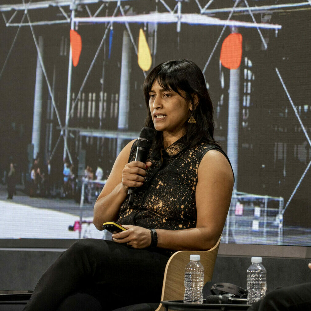 Η Shazna Nessa, Πρώην Παγκόσμια Επικεφαλής Οπτικού Υλικού της Wall Street Journa κρατά μικρόφωνο κι ένα presenter, καθώς κάθεται στην κεντρική σκηνή του Διεθνούς Δημοσιογραφικού Φόρουμ του iMEdD, δίπλα σε δύο μπουκαλάκια νερού. Παραδίδει την παρουσίασή της για το πάνελ «Ψηφιακός μετασχηματισμός: μια συνεχής διαδικασία». Πίσω της, μια μεγάλη οθόνη, όπου φαίνεται μια θαμπή διαφάνεια από την παρουσίασή της.
Λεζάντα: Φωτογραφία: Ρόννυ Σκεύης