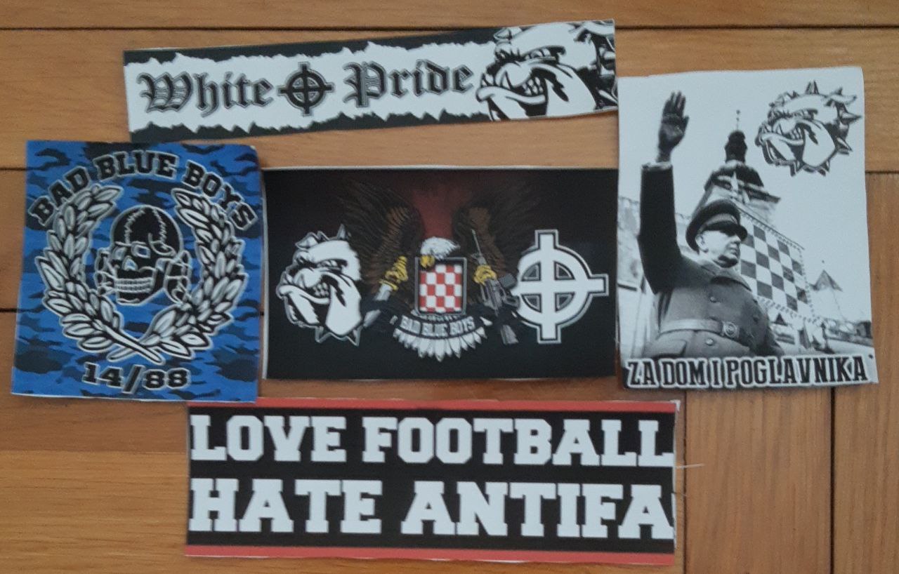 This picture shows five stickers related to the fan movement of the Croatian football team Dynamo Zagreb. On the top sticker is the phrase 'White Pride' and the far-right Celtic Cross symbol. Another sticker bears the phrase 'love football Hate antifa'. A third shows a skull with the words "Bad Blue Boys" above it and the numbers "14/88" below it. The fourth sticker, which is in the centre of the image, shows a dog's head, an eagle and the far-right Celtic Cross symbol, while "Bad Blue Boys" is written. The fifth sticker shows the Bad Blue Boys logo, and below it the founder of the Croatian fascist organisation Ustase, Ante Pavelic, waving in a manner reminiscent of the Nazi salute. 