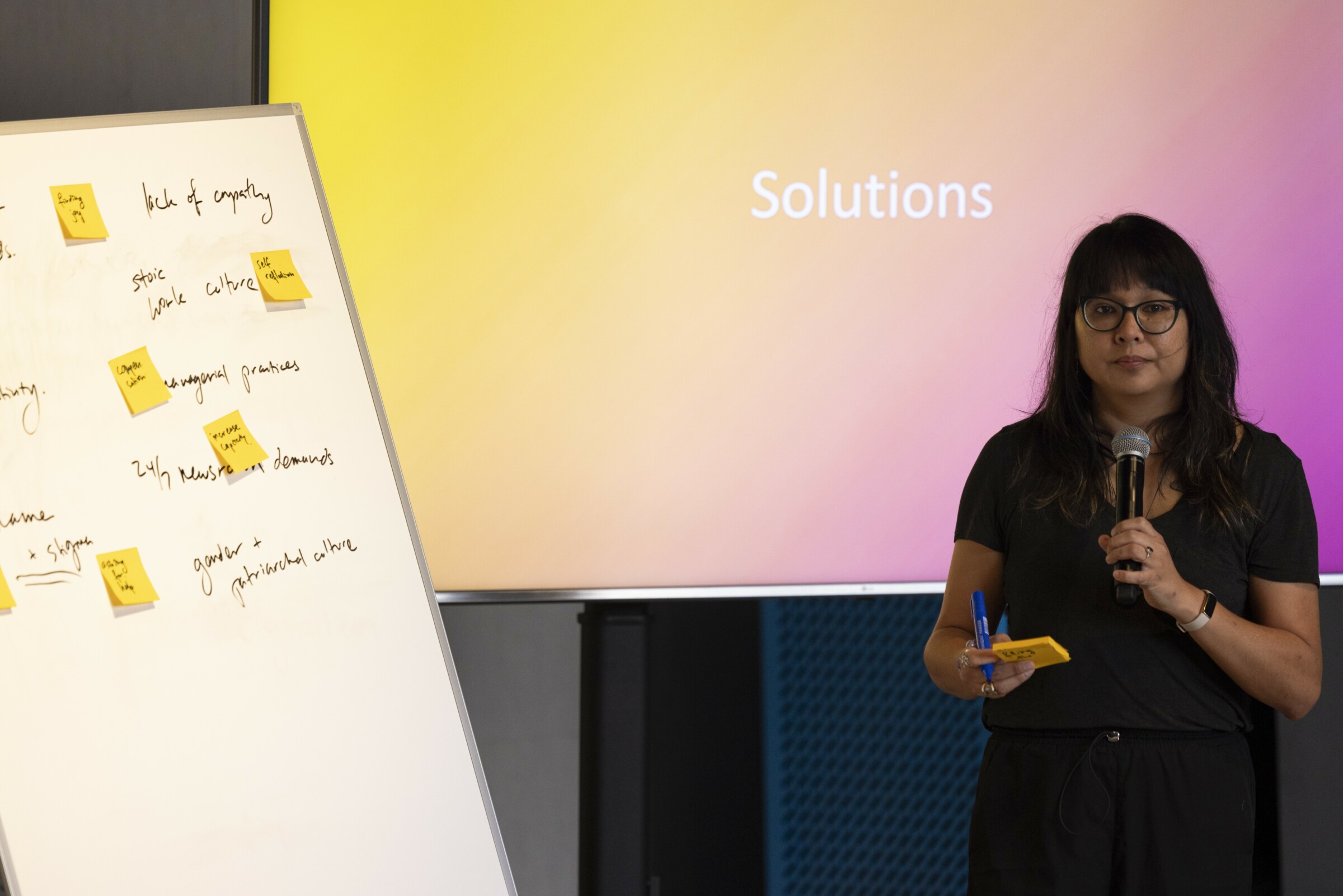 AX Mina (Ana) stands in front of a screen that displays the word "Solutions". To the left of the photo is a white board with notes and yellow post-its. She is holding a microphone in one hand and some post-its in the other. The audience is shown in the foreground, out of focus. She wears glasses and is dressed in simple black clothing. Photo: Alex Grymanis