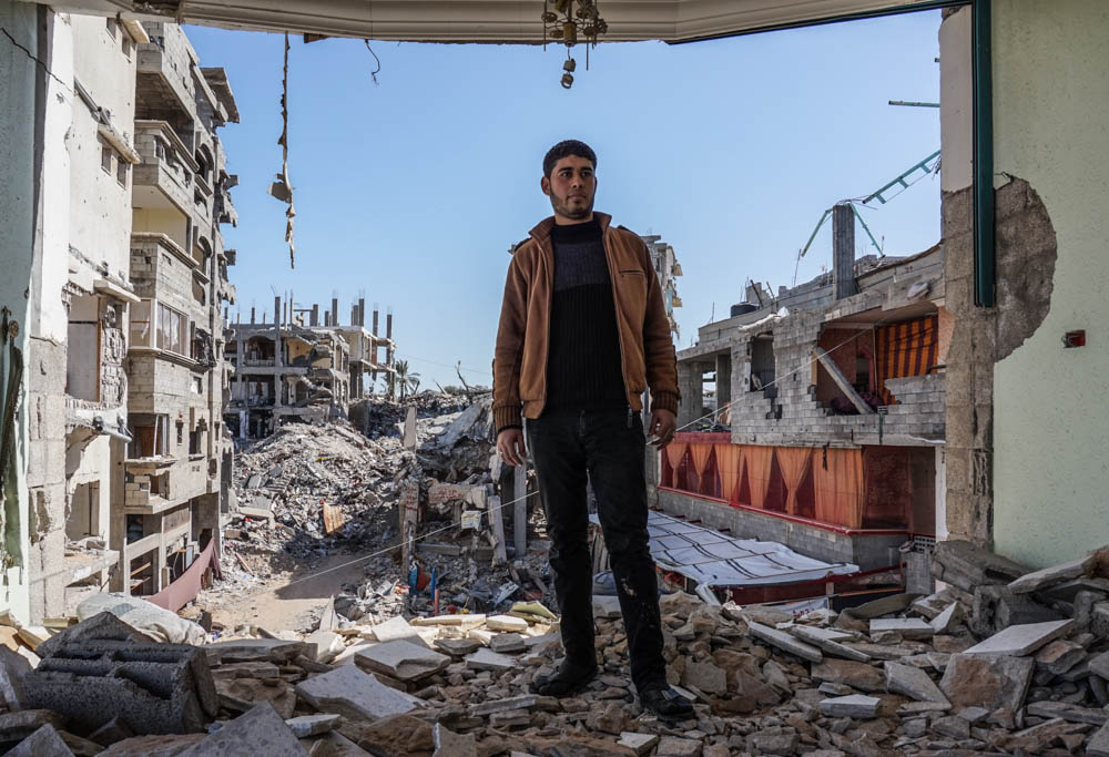 The picture shows a resident of the Gaza Strip, specifically the Shujayea area, standing in his destroyed house. In the background, many other destroyed buildings can be seen. 