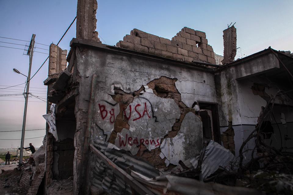 Η εικόνα δείχνει ένα βομβαρδισμένο και κατεστραμμένο σπίτι στη Λωρίδα της Γάζας, το 2014. Στον μπροστινό τοίχο αναγράφεται η φράση "By USA Weapons" (από αμερικανικά όπλα).