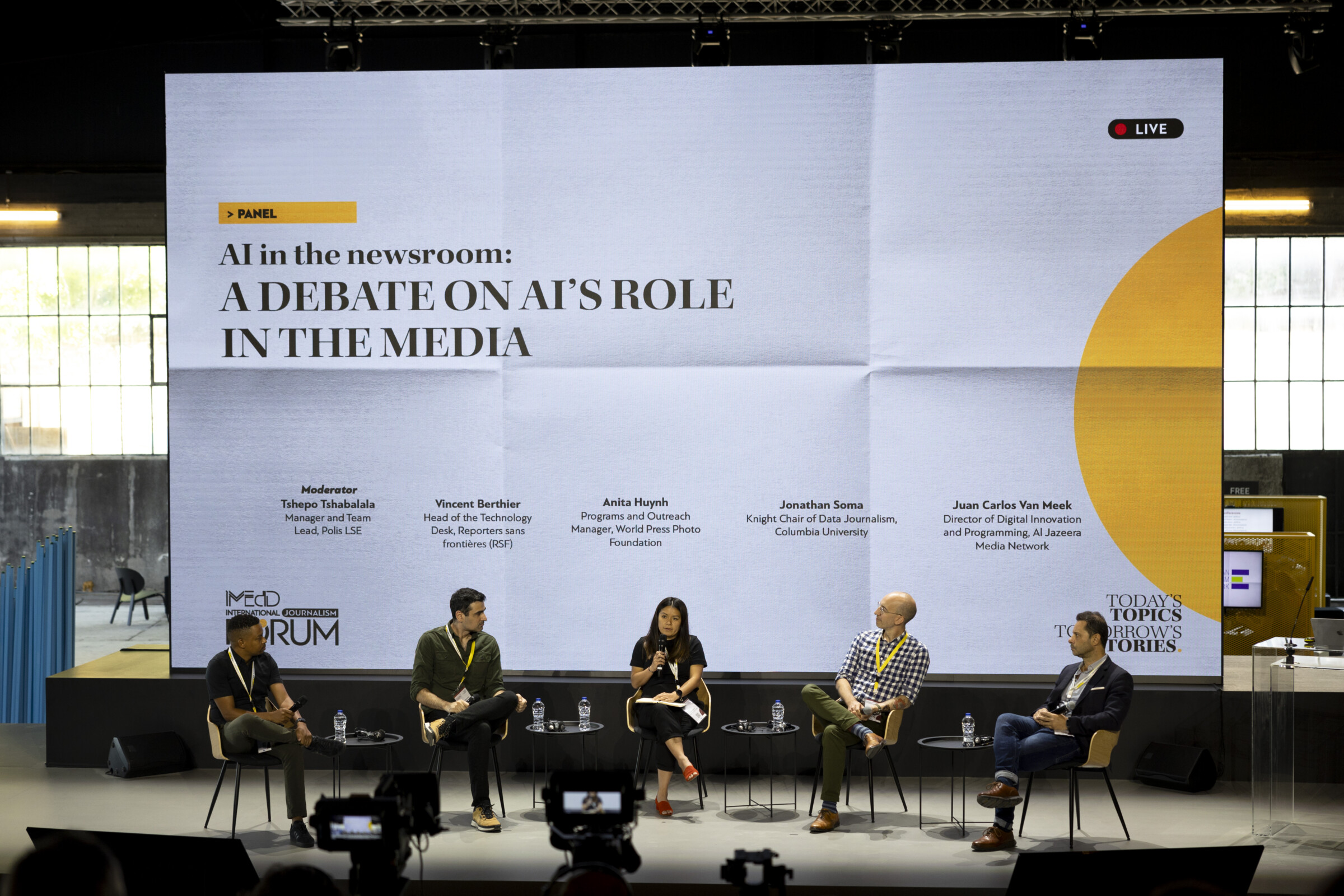 Σε μια σκηνή πέντε άτομα κάθονται μπροστά από ένα μεγάλο λευκό και κίτρινο πανάκι που γράφει "AI in the Newsroom: Μια συζήτηση για το ρόλο της τεχνητής νοημοσύνης στα μέσα ενημέρωσης". Η γυναίκα στο κέντρο, ντυμένη στα μαύρα, κρατάει το μικρόφωνο. Οι υπόλοιποι τέσσερις άνδρες του πάνελ την ακούνε όλοι μαζί.