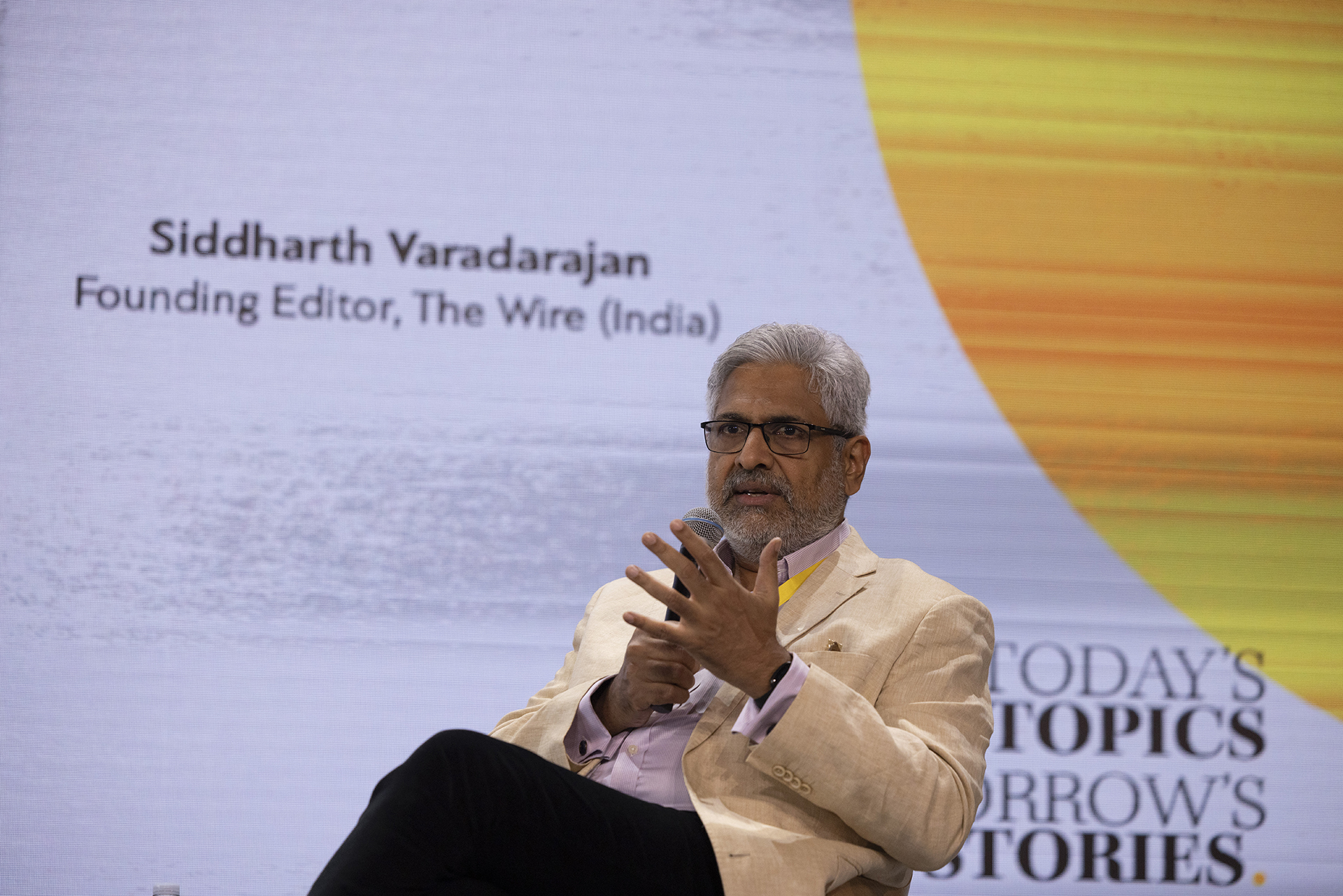 Siddharth Varadarajan, Founding Editor at The Wire, at the panel “Sustainability models in an era of digital revolution” during  iMEdD International Journalism Forum 2023. He sits on a chair on stage holding a hand-held  microphone and raising his hand as a sign of determination and intensity. On the background screen stands his name and title.
