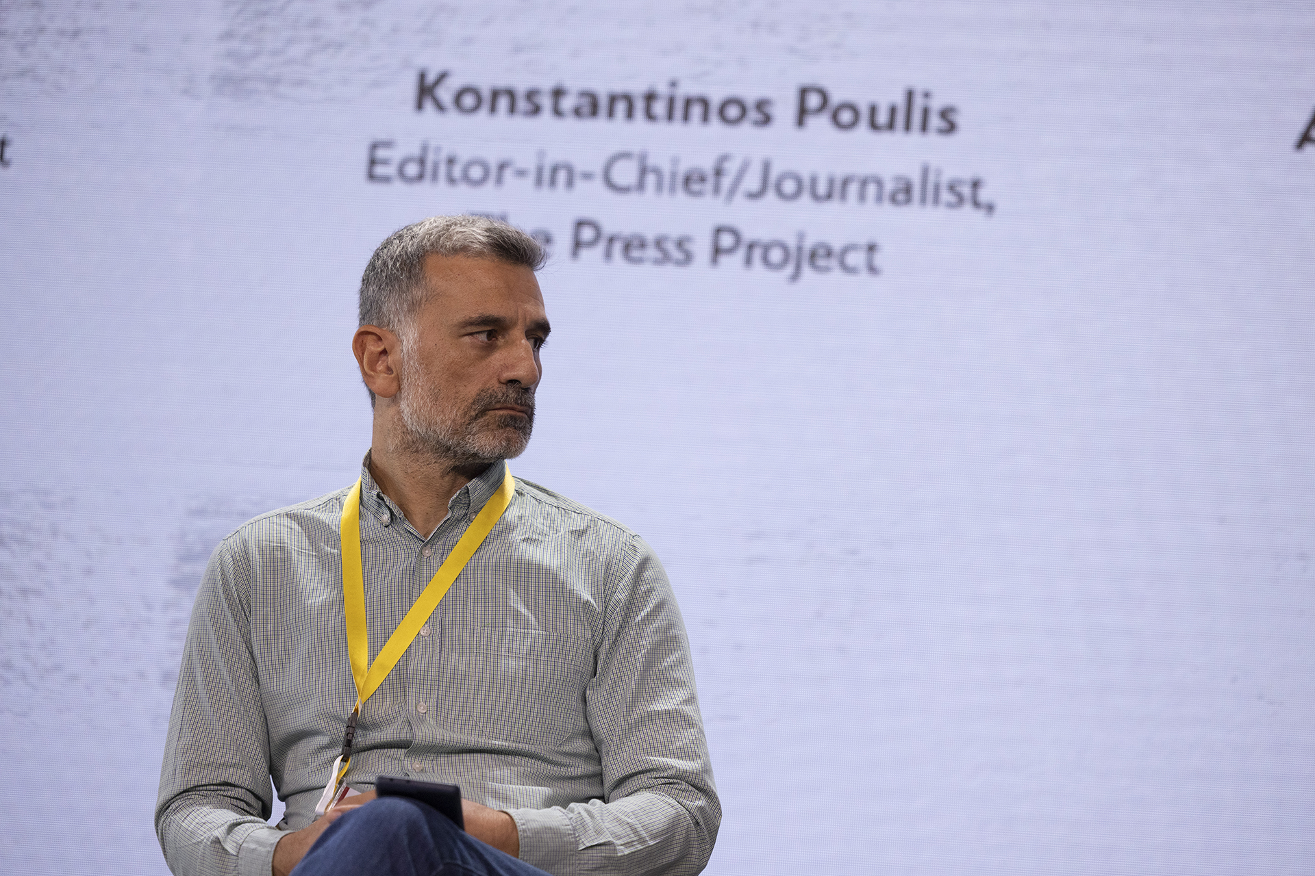 Konstantinos Poulis, Editor-in-Chief at The Press Project, at the panel “Sustainability models in an era of digital revolution” during  iMEdD International Journalism Forum 2023. He is sitting on a chair looking reflective on his left side. On the background screen stands his name and title.