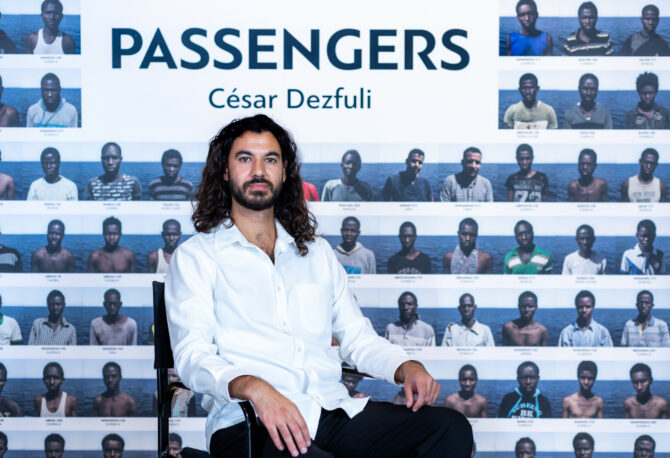 Αυτή η εικόνα, η οποία τραβήχτηκε στο πλαίσιο του Διεθνούς Φόρουμ Δημοσιογραφίας του iMEdD, δείχνει τον Ισπανό-Ιρανό φωτοδημοσιογράφο Σεζάρ Ντεζφούλι να ποζάρει μπροστά από το κεντρικό banner της φωτογραφικής έκθεσης του project του με τίτλο "Passengers"