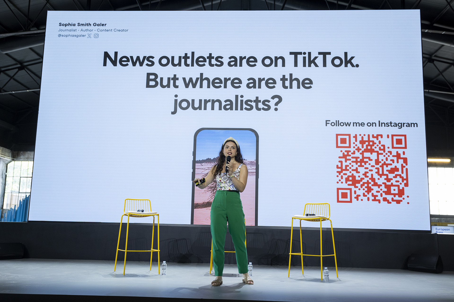 Η Sophia Smith Galler στην κεντρική ομιλία της "Τα ειδησεογραφικά brands είναι στο TikTok - αλλά πού είναι οι δημοσιογράφοι;" στέκεται στο κέντρο της σκηνής φορώντας ένα σκουρο πράσινο παντελόνι, μια μπλούζα με λουλούδια και κορδέλα για τα μαλλιά. Στην οθόνη πίσω της εμφανίζεται ο τίτλος της ομιλίας της και ένας κωδικός QR που οδηγεί στον λογαριασμό της στο Instagram.
