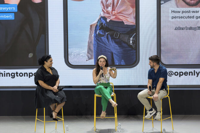 The three panelists of "Storytelling in vertical video: How news can use TikTok (and all the apps that are copying it)" sitting on yellow stools on the stage of iMEdD International Journalism Forum 2023. Sophia Smith Galler is standing at the center of the stage and speaking in a hand-held microphone, while Carmella Boykin on her right and Enrique Anarte Lazo on her left are looking at her. They are all smiling and excited about the discussion. The background screen shows screenshots from some of their most successful TikTok videos.