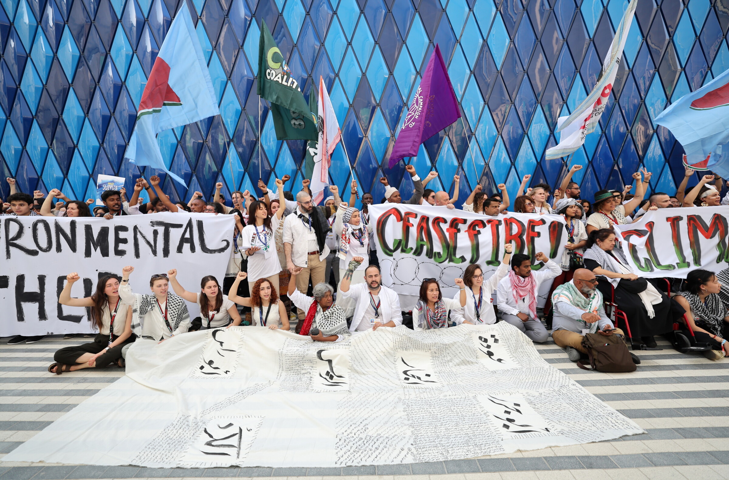 Activists protesting in favour of the Palestinian people during COP 28 at Expo City Dubai. Some are sat and some are stood holding flags with the watermelon symbol of Palestinian solidarity and banners reading "Environmental Apartheid", and "Ceasefire", among others.  Photo: Ali Haider, EPA, 03/12/2023