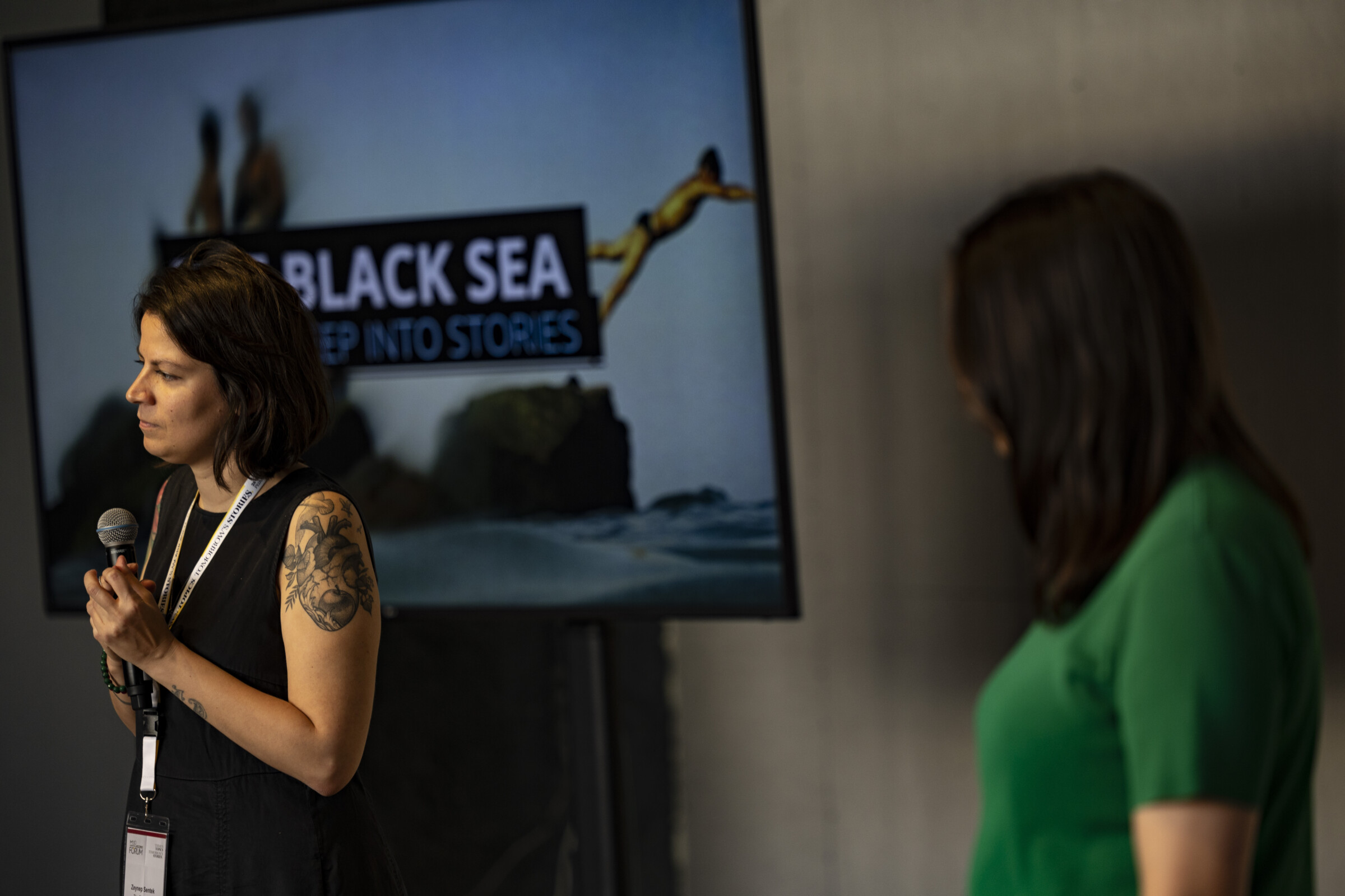 Δύο γυναίκες παρουσιάζουν ένα workshop στο πλαίσιο του Διεθνούς Φόρουμ Δημοσιογραφίας του iMEdD. Η μία από αυτές, η Zeynep Sentek κρατάει ένα μικρόφωνο, ενώ από πίσω της υπάρχει μια οθόνη με το λογότυπο του οργανισμού THE BLACK SEA.