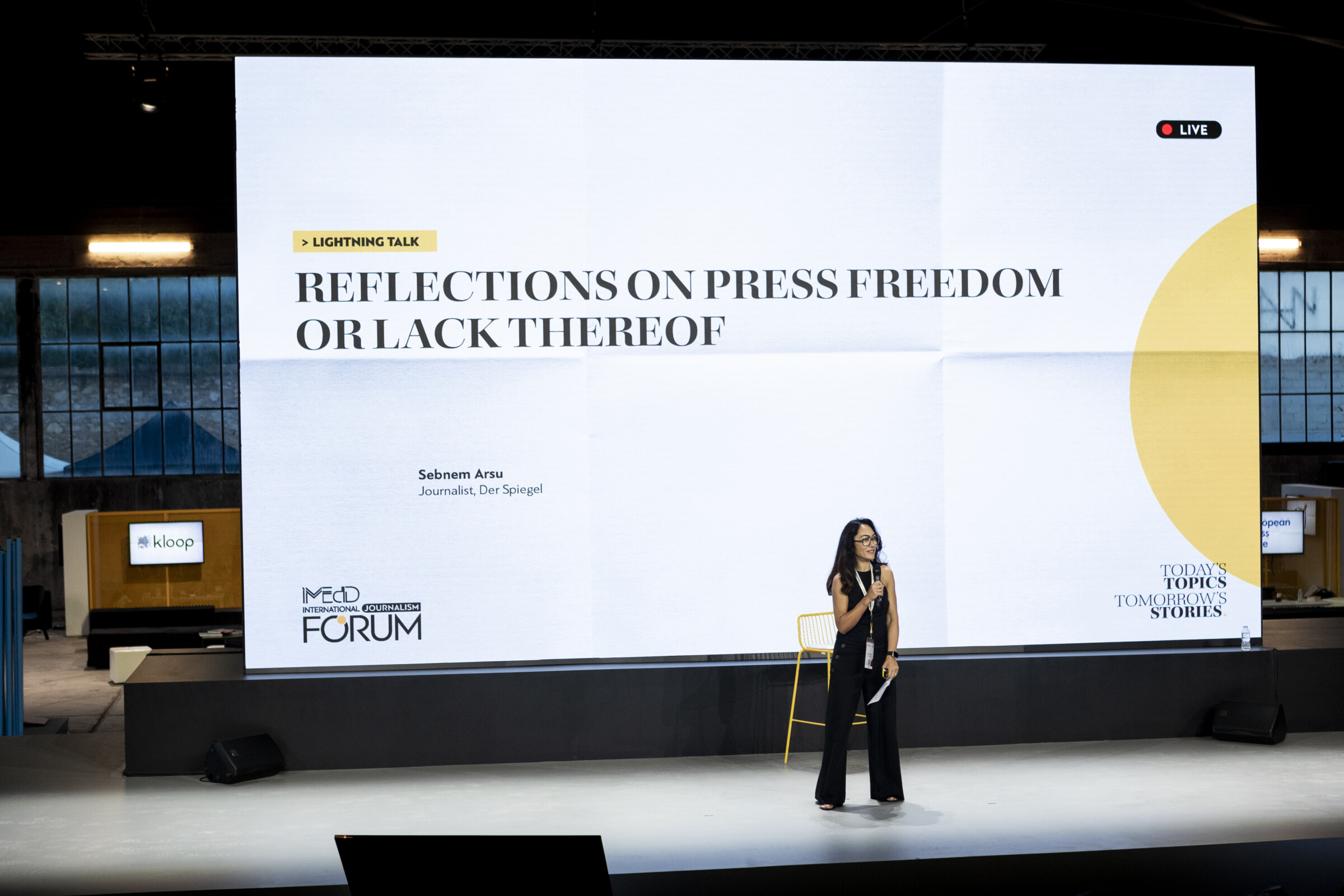 Turkish journalist Sebnem Arsu stands on the main stage of the iMEdD International Journalism Forum. She is holding a microphone and behind her is a large screen with the title of the panel she is participating in , entitled "Reflections on press freedom or lack thereof".