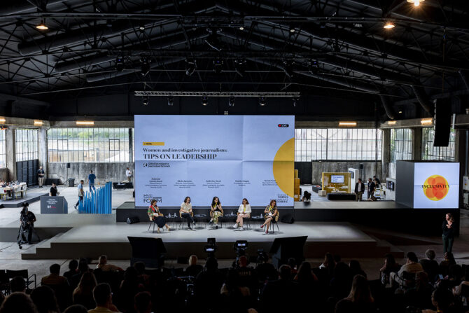Long shot of the panel discussion "Women and investigative journalism: Tips on leadership". The four panelists sit on chairs next to each other. In the background stands the screen with the title of the panel, as well as the names and titles of the panelists. Except for the stage, we can also see the media village behind it and the audience sitting in the first rows.