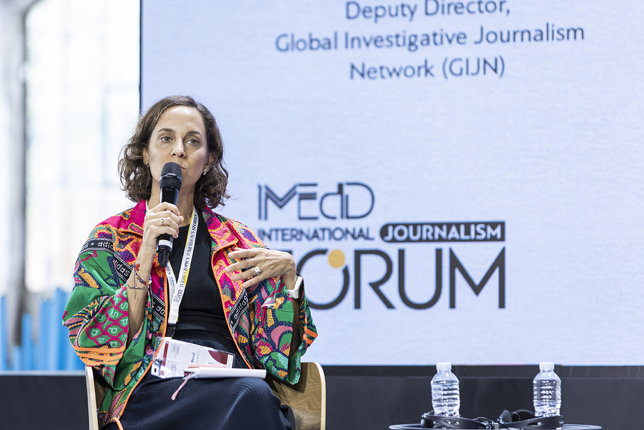 Gabriela Manuli on the stage of iMEdD International Journalism Forum 2023. She is holding a hand-held microphone in her right hand, while she is moving her left one. She is wearing a colourful jacket over a black top and a pair of black trousers.