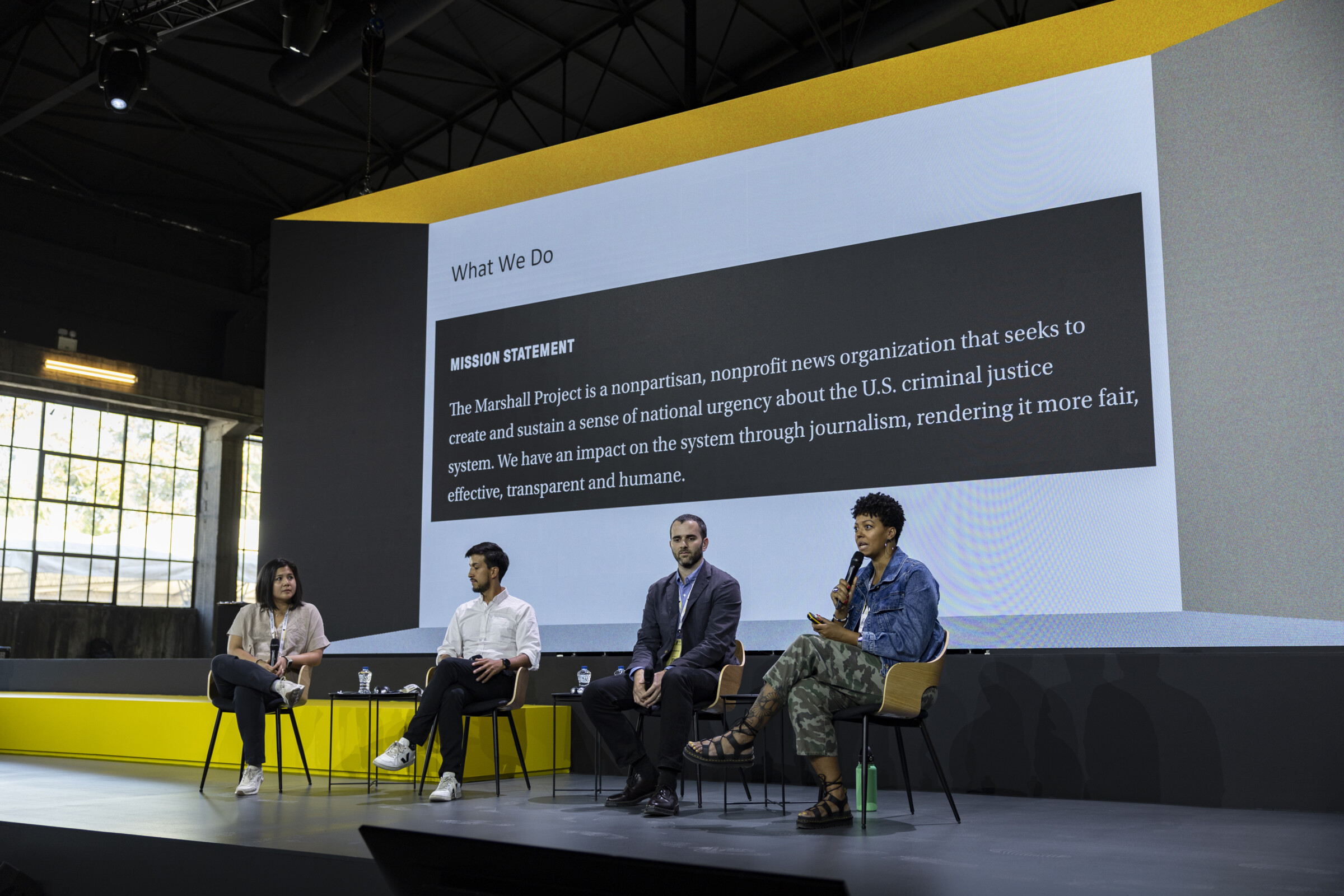 A wide shot of a panel discussion on building communities at the iMEdD's International Journalism Forum 2024. Four panelists sit on stage, each with microphones in their hands. A screen behind them displays the panel title and conference logo. The panelists are mid-discussion, with one person gesturing while speaking. 