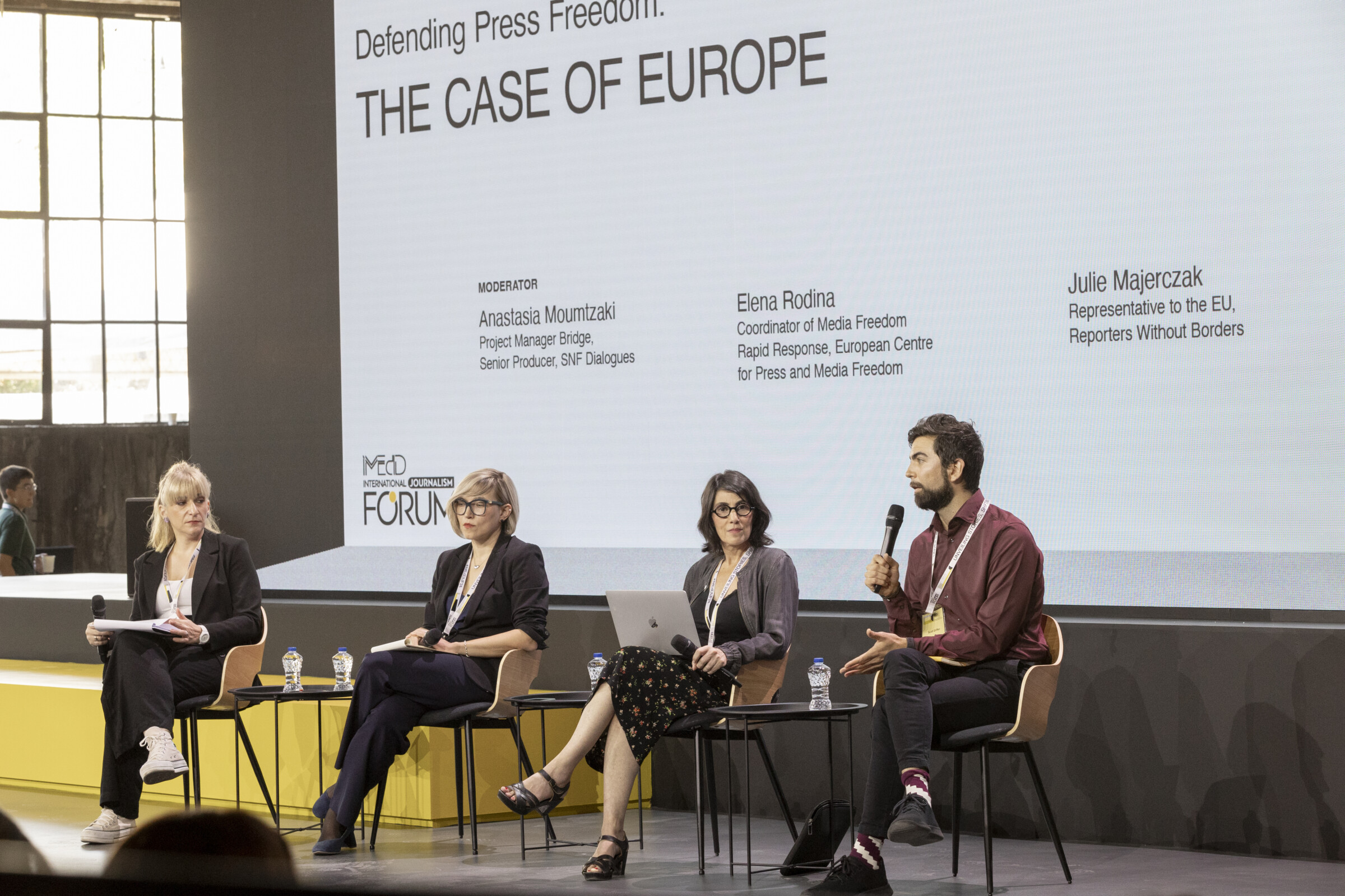 Anastasia Moumtzaki, Elena Rodina, Julie Majerczak, and Scott Griffen sit onstage. Scott Griffen speaks to the microphone. 