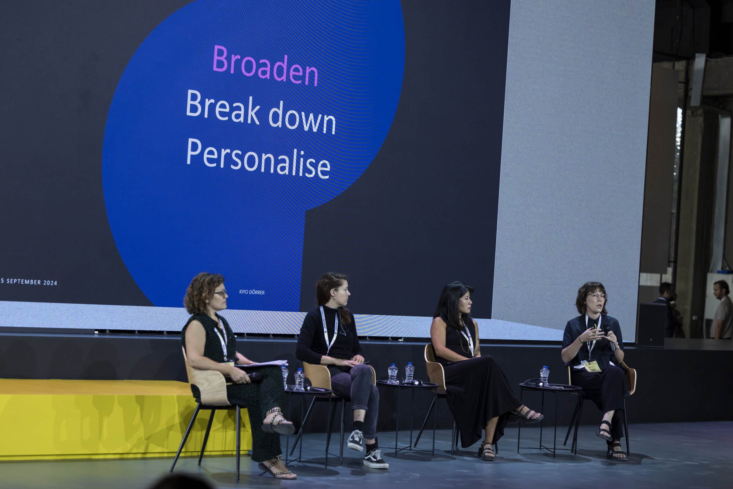 Four female panelists seated on stage, one holding a microphone, engaging in a discussion. From right to left, Vera Penêda, Ingrid Gercama, Joi Lee, and Kiyo Dörrer.