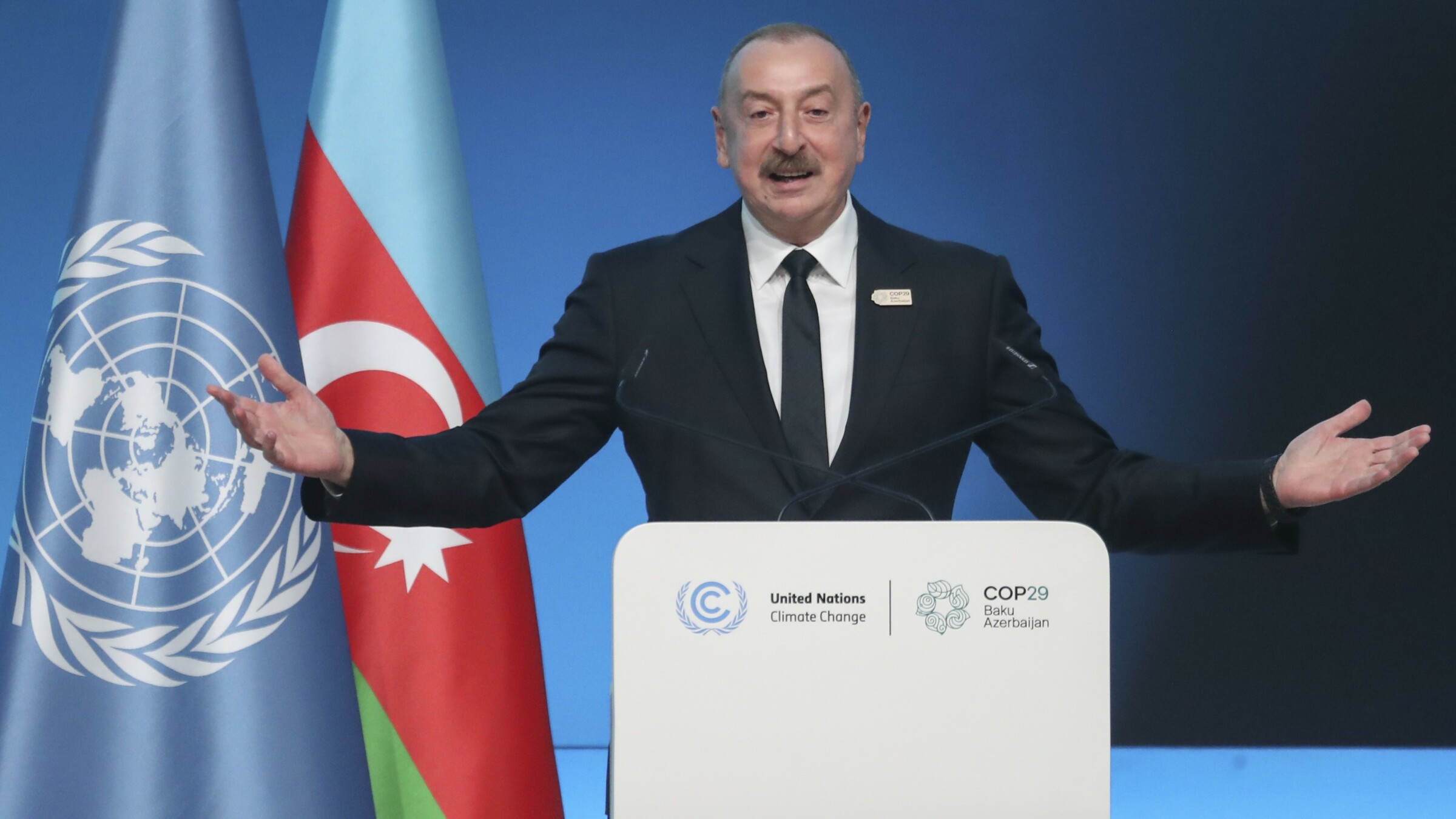 Azerbaijan's President, Ilham Aliyev, addressing the attendees at COP29 (12 November 2024). He has his arms wide open, standing behind a podium with the "United Nations Climate Change" and "COP 29 Baku Azerbaijan" logos on it. Behind him, a United Nations flag and an Azerbaijan flag. 