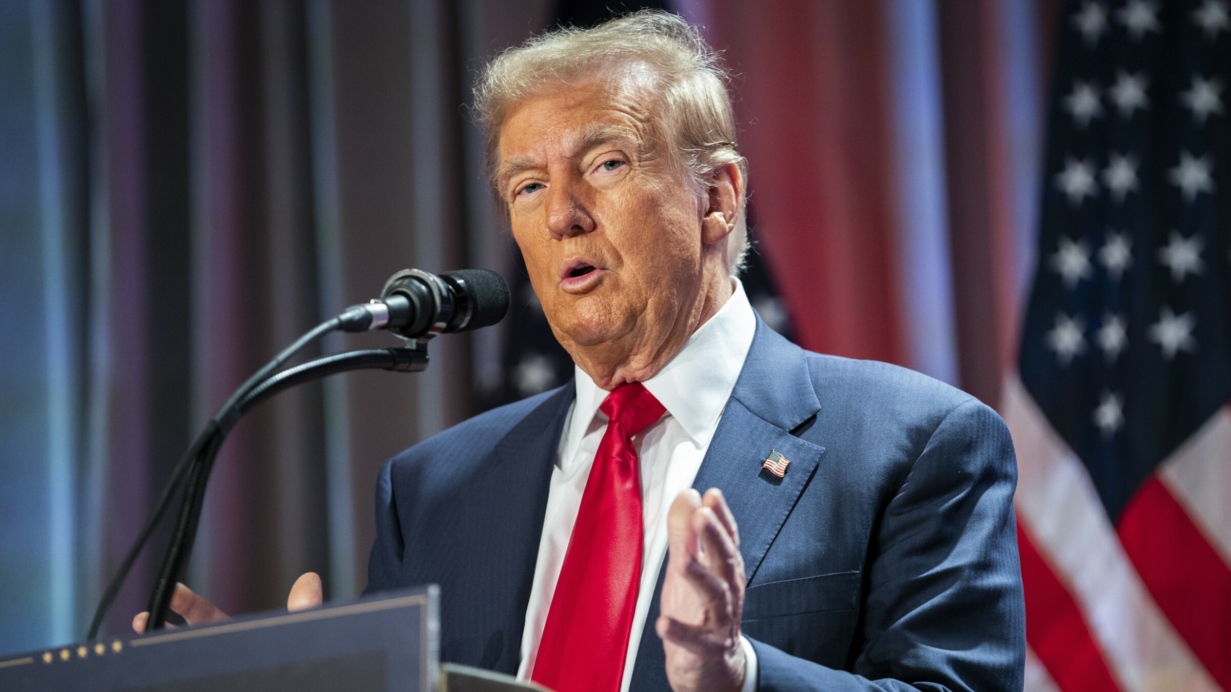 President-elect, Donald Trump, during a meeting with House Republicans in Washington (13 November 2024). He is speaking into a microphone, standing behind a podium with his hands open. Behind him, two American flags.