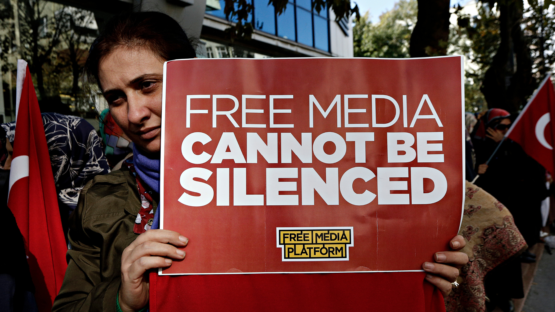 A female journalist is holding a red sign that reads Free Media Cannot be Silenced