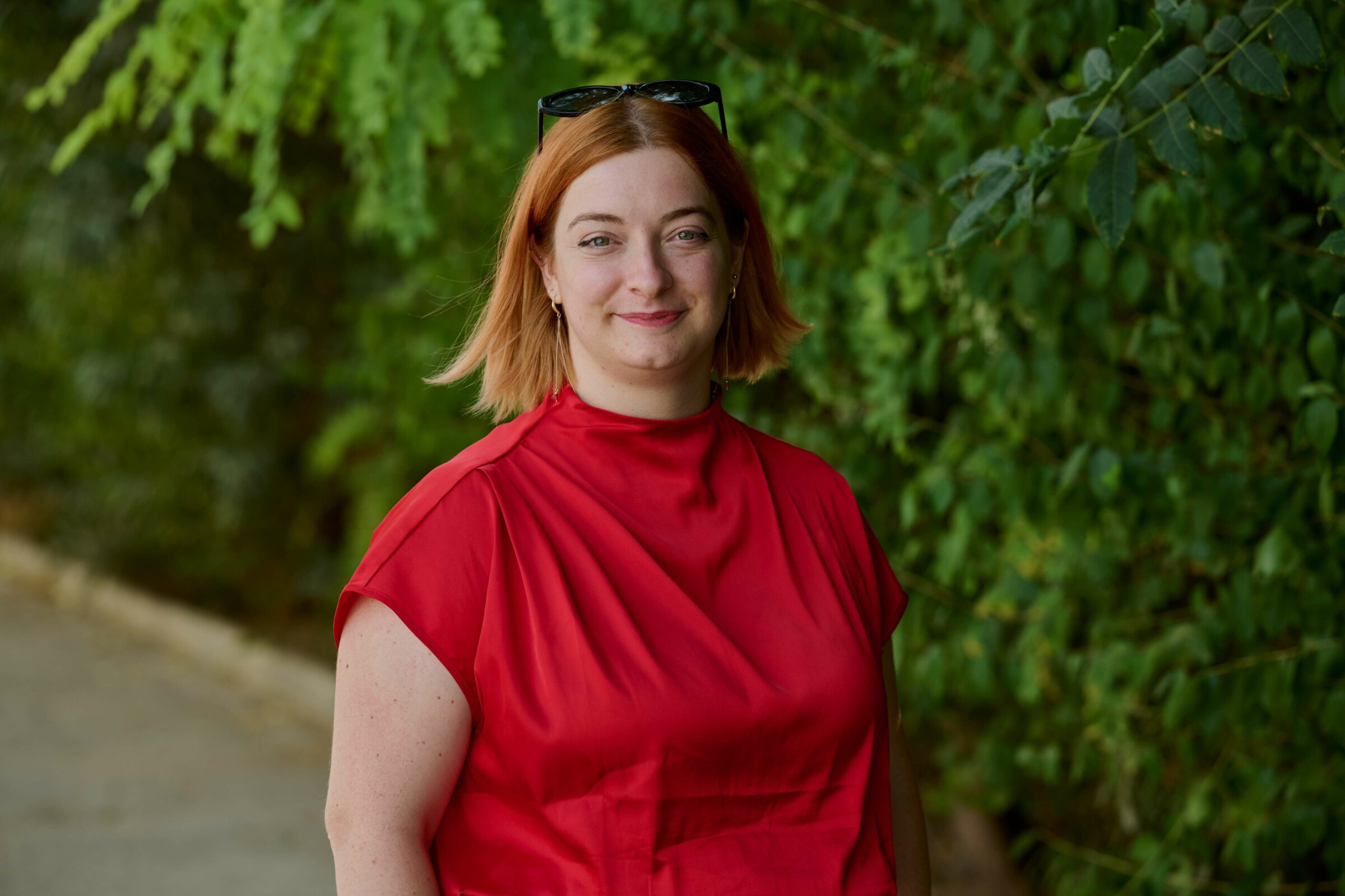 Erika Marzano, audience development manager at Deutsche Welle, photographed by Petros Toufexis. She has ginger short hair and blue eyes. She is wearing a read blouse and has glasses on top of her head. She is stood still with a green plant backdrop. She is smiling.
