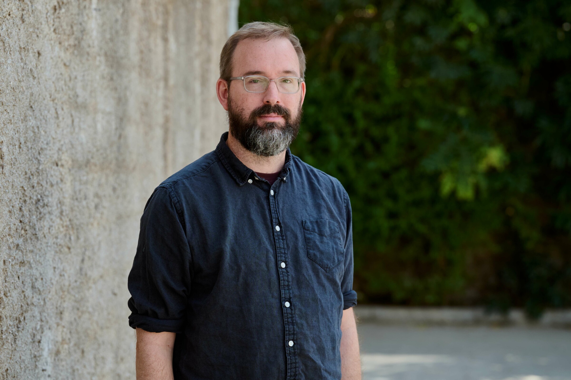 Dr. Daniel Klug, Senior Multimedia Production lecturer at the University of Applied Sciences of the Grisons, photographed by Petros Toufexis. He has black and grey hair, a beard and brown eyes. He is wearing a dark blue shirt and glasses. He is stood still, with a gray wall and some green plants as his background. He is slightly smiling.